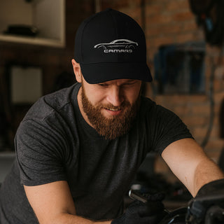 A rugged bearded man wearing a black Camaro embroidered hat works on a car in a garage, surrounded by tools and equipment.