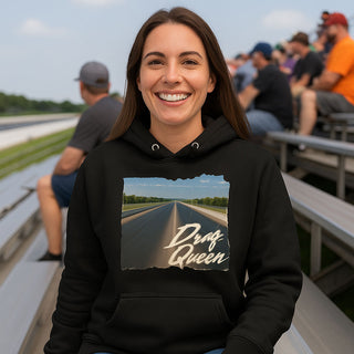 A smiling woman wearing a black “Drag Queen” hoodie sits on the bleachers at a race track, with blurred spectators in the background.