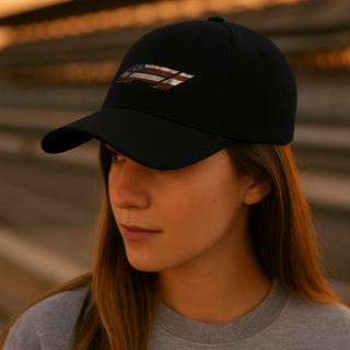 Person wearing a black embroidered dad hat with distressed American flag Formula 1 logo, standing in front of a racetrack grandstand at sunset — Geaux Big motorsport lifestyle photo.