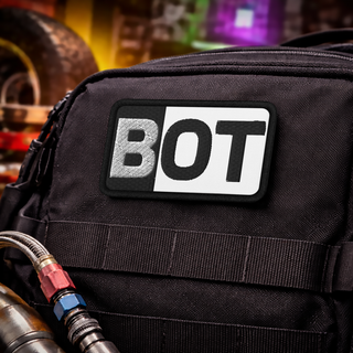 Black gear bag with a black-and-white embroidered BOT patch attached on the front, photographed in an automotive workshop setting with blurred Mardi Gras-inspired purple, green, and gold lighting in the background.